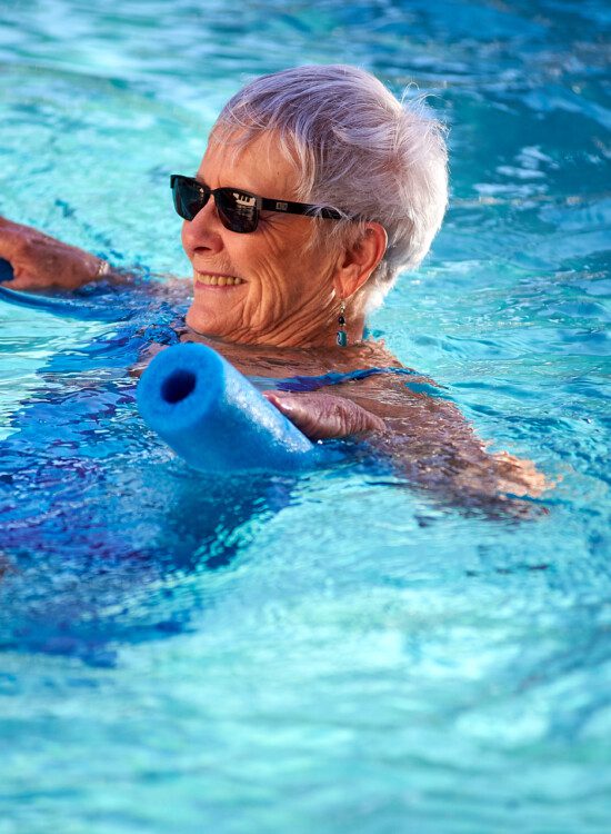 A senior women in a pool swimming with a pool noodle