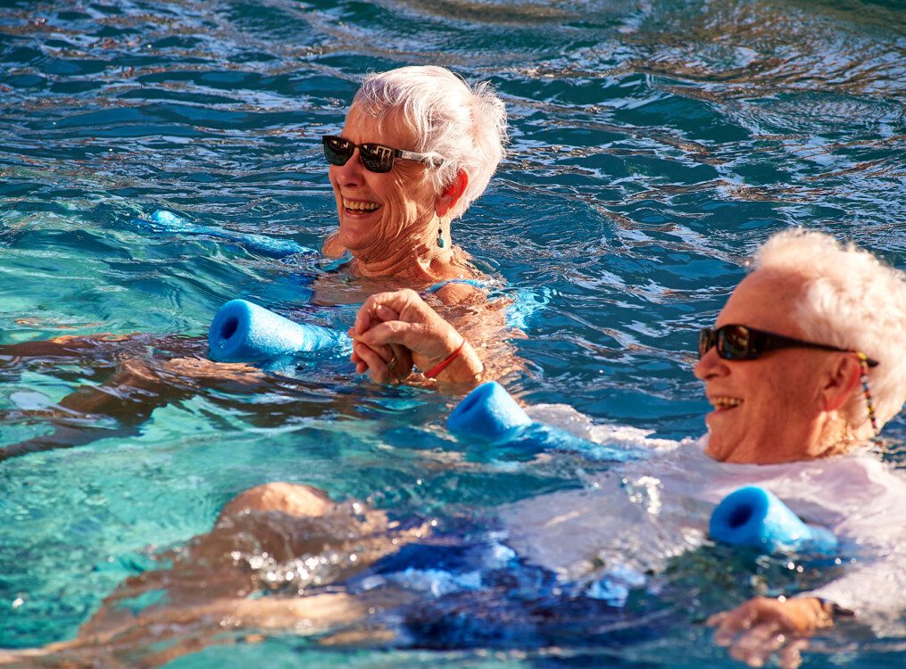 Two seniors in a pool swimming with a pool noodle